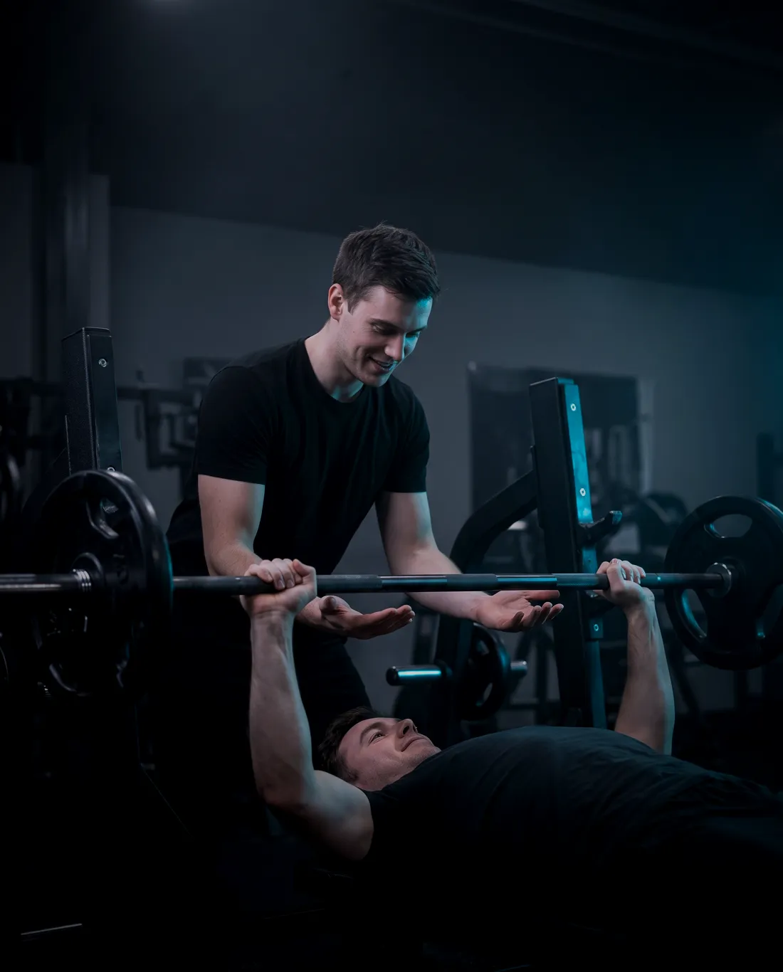 Dva lidé u bench pressu: jeden zvedá, druhý ho jistí a usmívá se